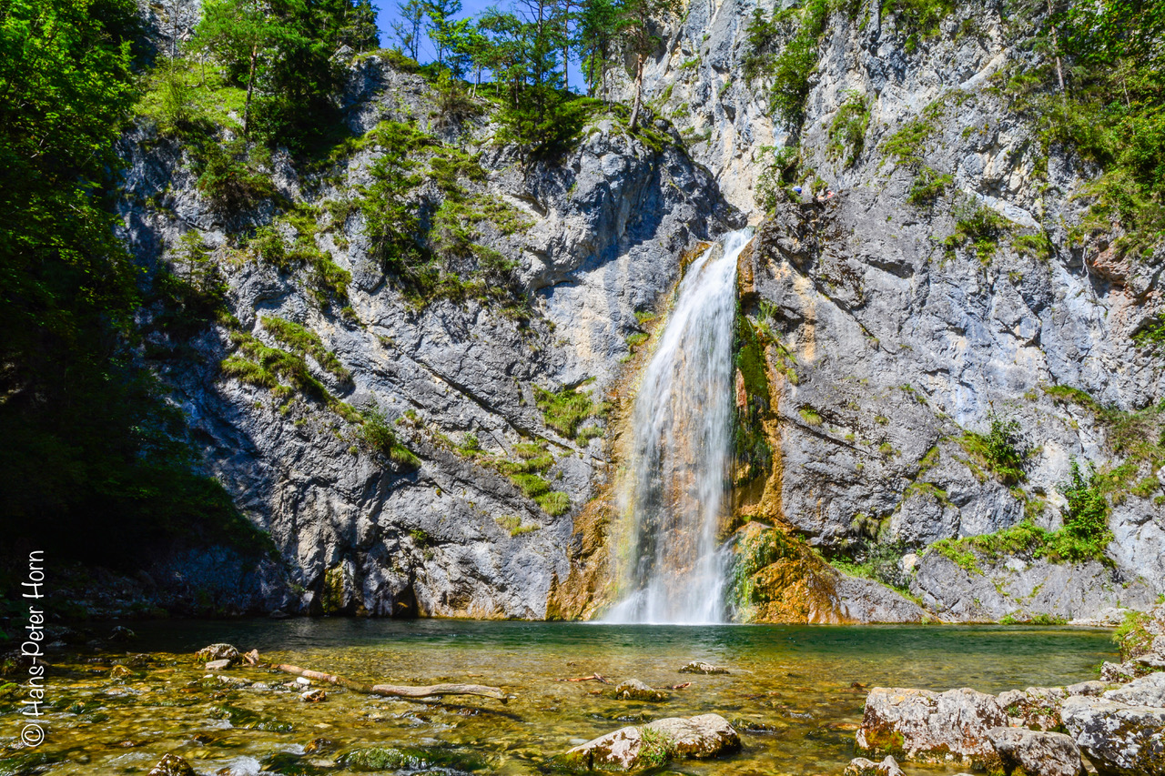 Steinwand mit Büschen bewachsen, Wasserfall fällt in der Mitte herunter, mündet in klarem, schimmerndem Wasser,